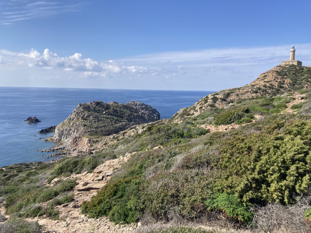 Il panorama del faro di Capo Sandalo sull'isola di San Pietro. Cielo blu con delle nuvole, il mare blu, costiera ricca di macchia mediterranea del colore verde intenso.